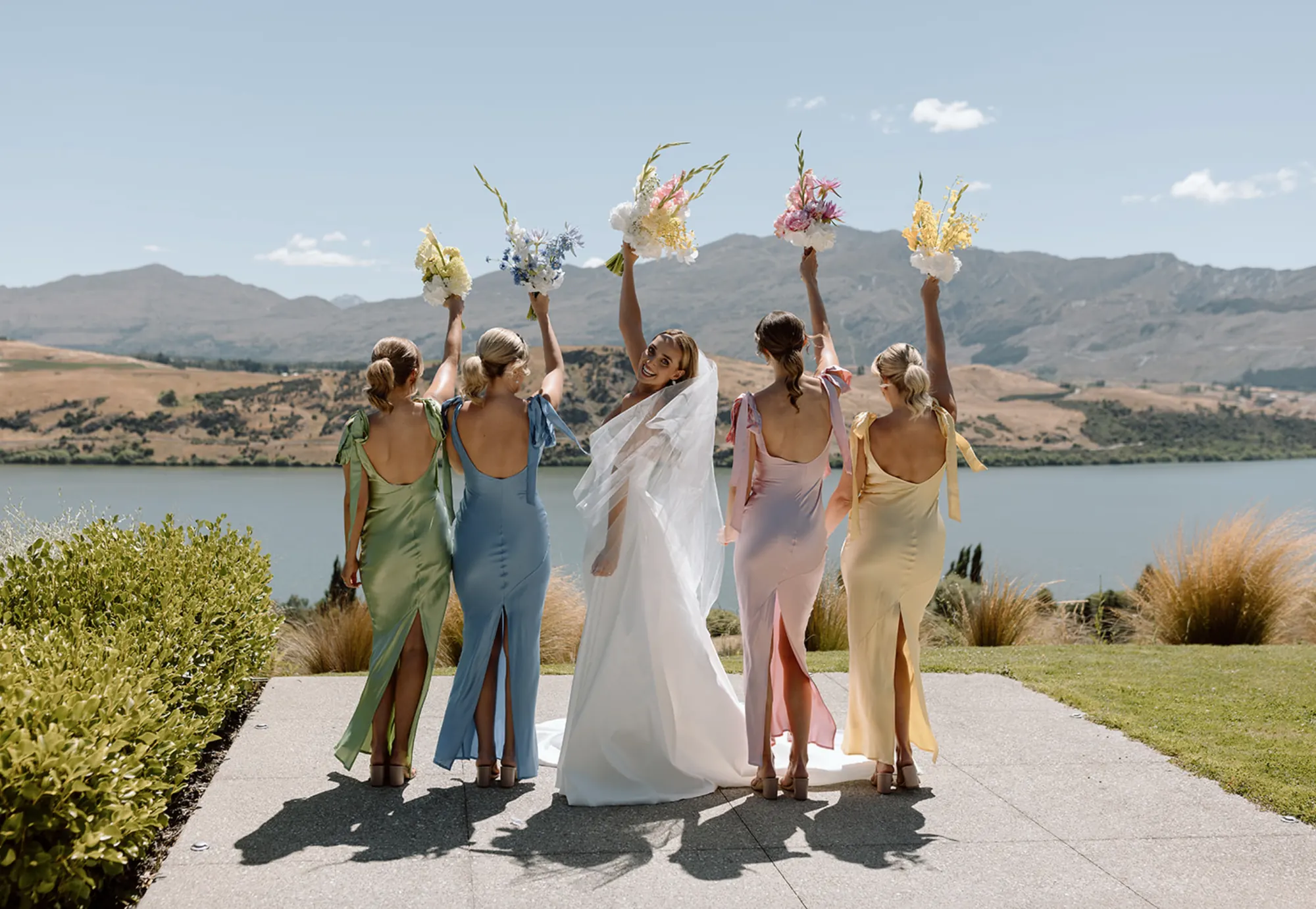A bride and her bridesmaids in pastel satin raising their bouquets at a New Zealand wedding Stacey styled